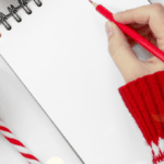 A woman's hand with red pencil surrounded by holiday artifacts, Santa hat, etc. preparing to make a list on a white sheet of paper