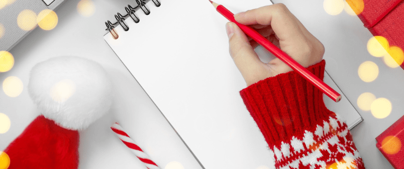 A woman's hand with red pencil surrounded by holiday artifacts, Santa hat, etc. preparing to make a list on a white sheet of paper