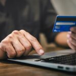 a hand typing on a laptop with another hand holding a credit card -- shopping online